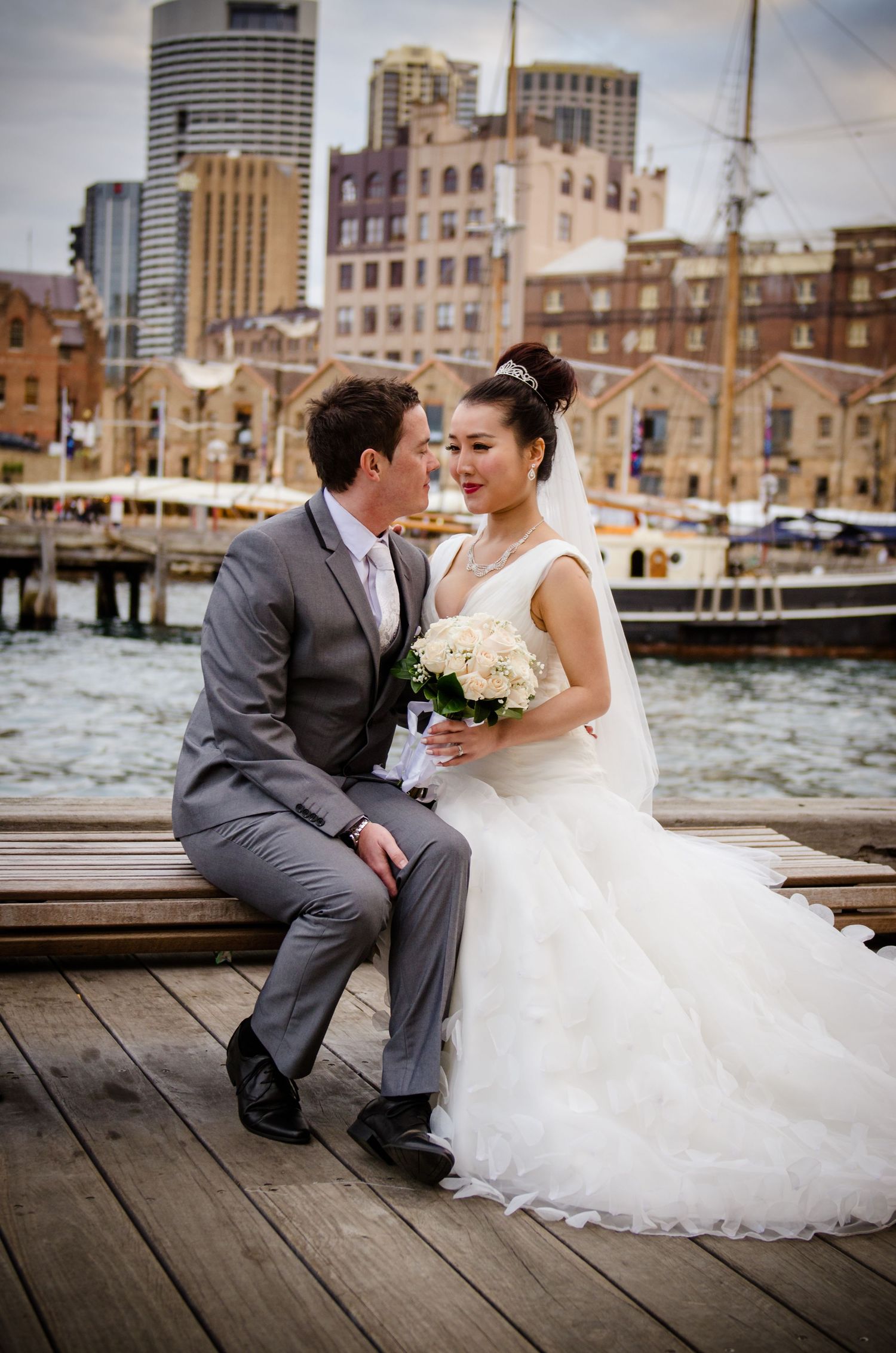 Wedding couple seated on wooden dock with urban waterfront backdrop.