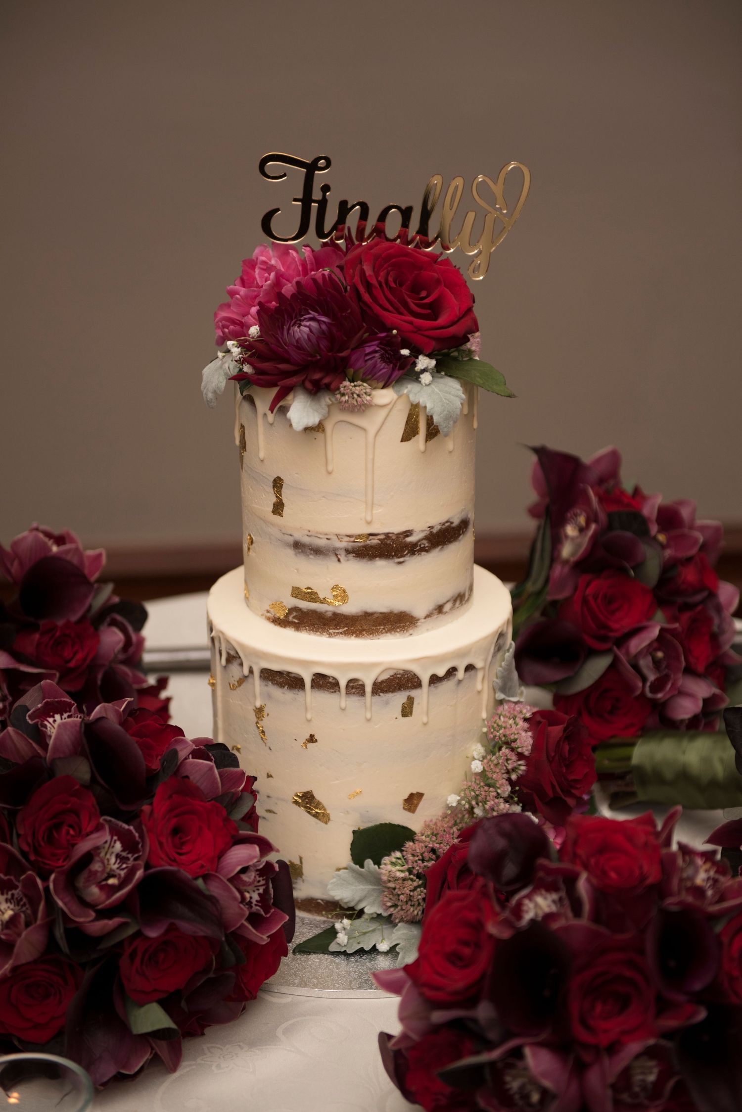 Elegant white wedding cake with gold drips decorated with deep burgundy roses and floral arrangements.