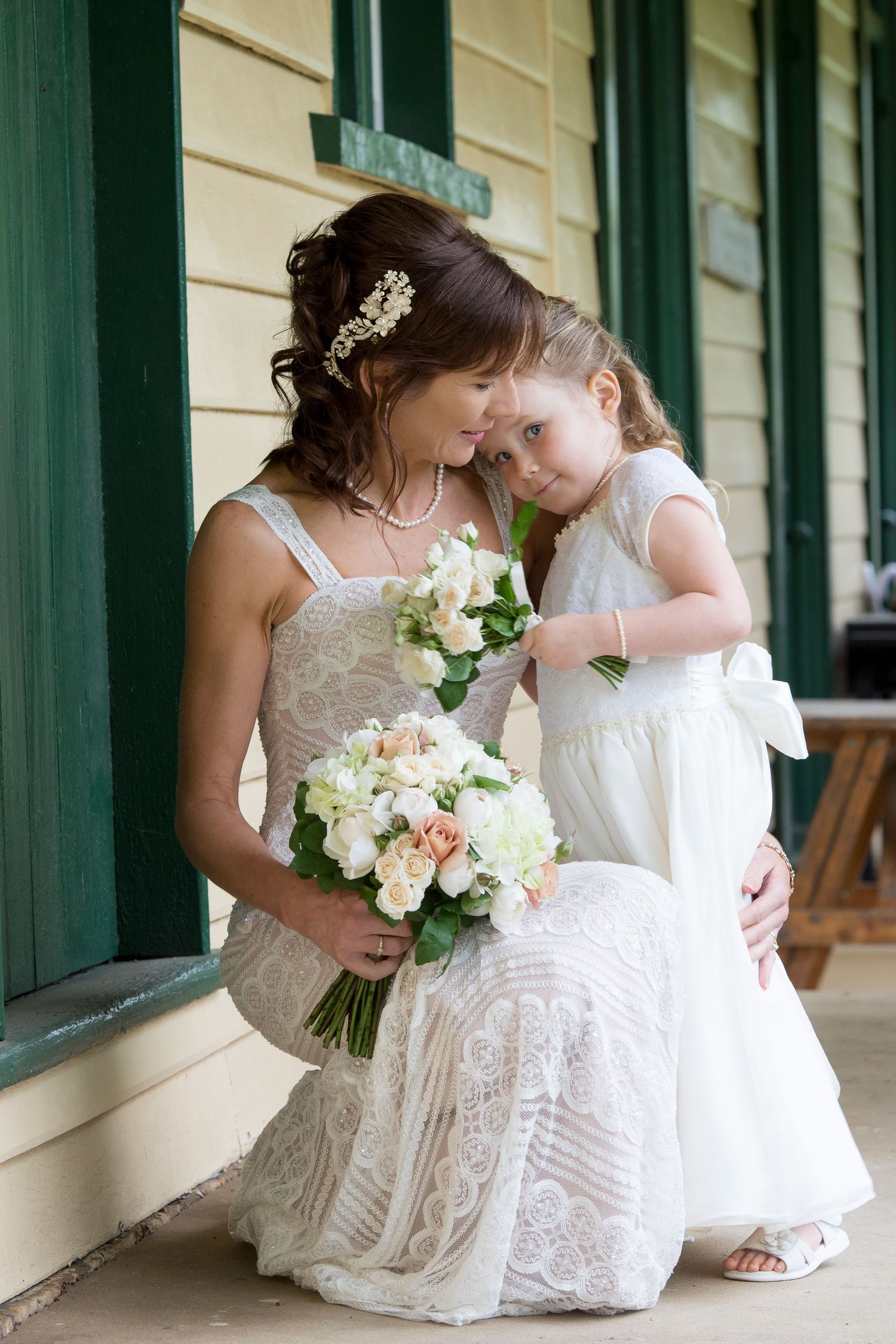 Bride in lace wedding dress shares tender moment with flower girl in white dress on covered porch.