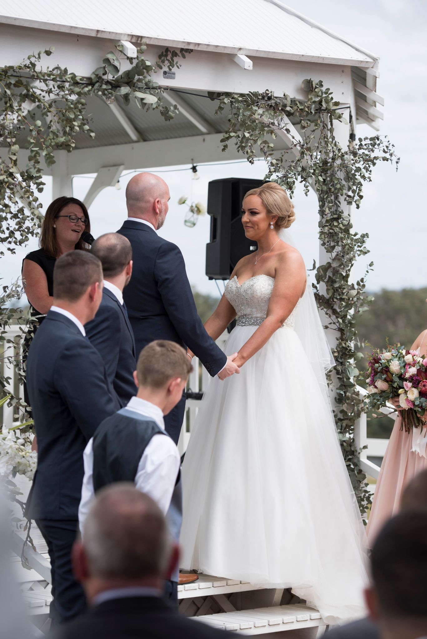 Wedding ceremony under a white gazebo with guests watching as couple exchanges vows during outdoor celebration.