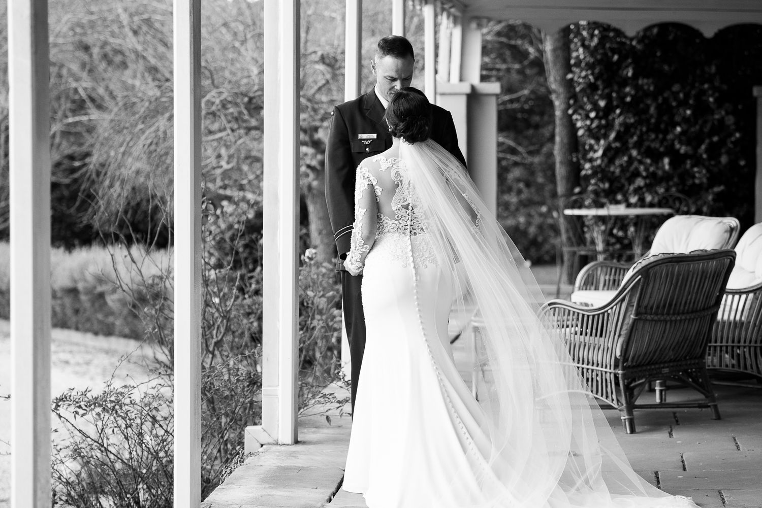 A bride in a flowing white gown and veil sharing a moment with groom on front porch.