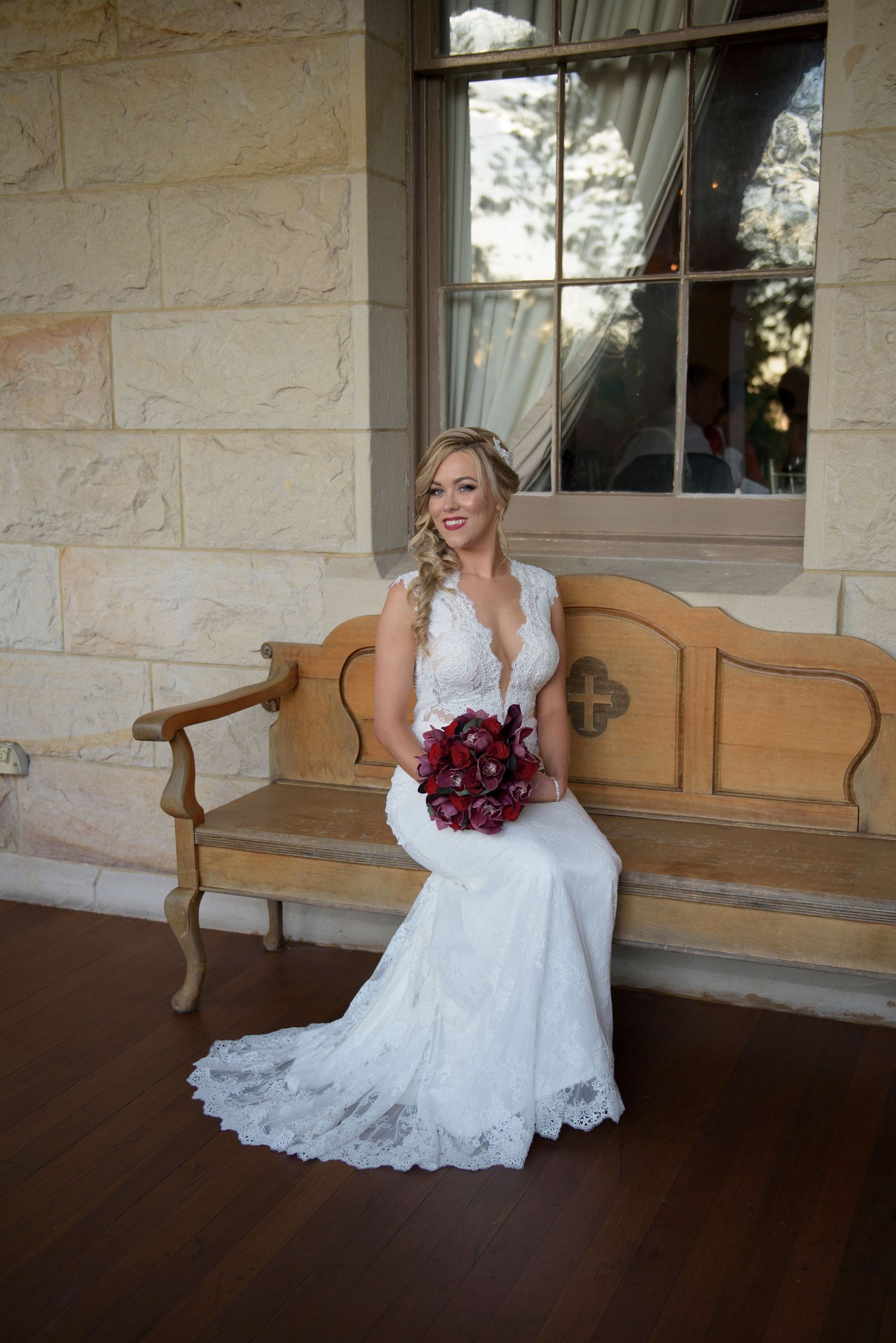 Bride in lace dress sits on wooden bench holding red bouquet at stone building.