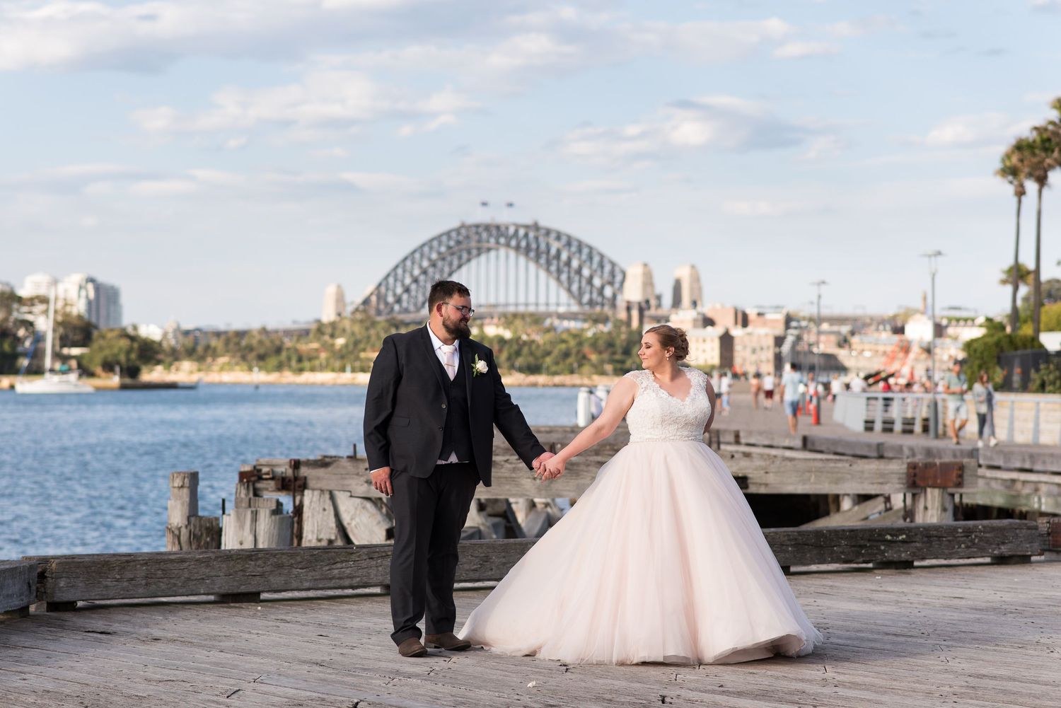 Bride and groom walks along a waterfront promenade during a sunny afternoon with Sydney Harbour Bridge in background.