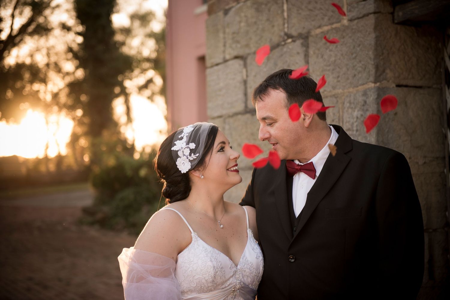 Romantic couple shares tender moment at sunset against stone wall with rose petals falling in golden light.