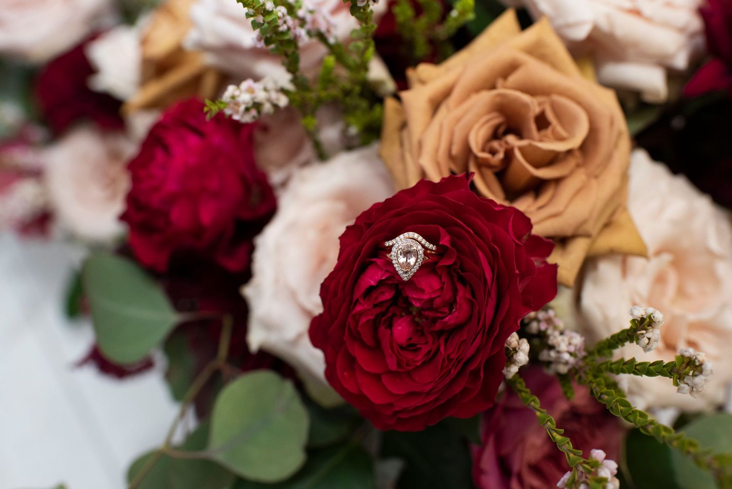 A wedding ring nestled in deep red and peach roses with delicate baby's breath accents.