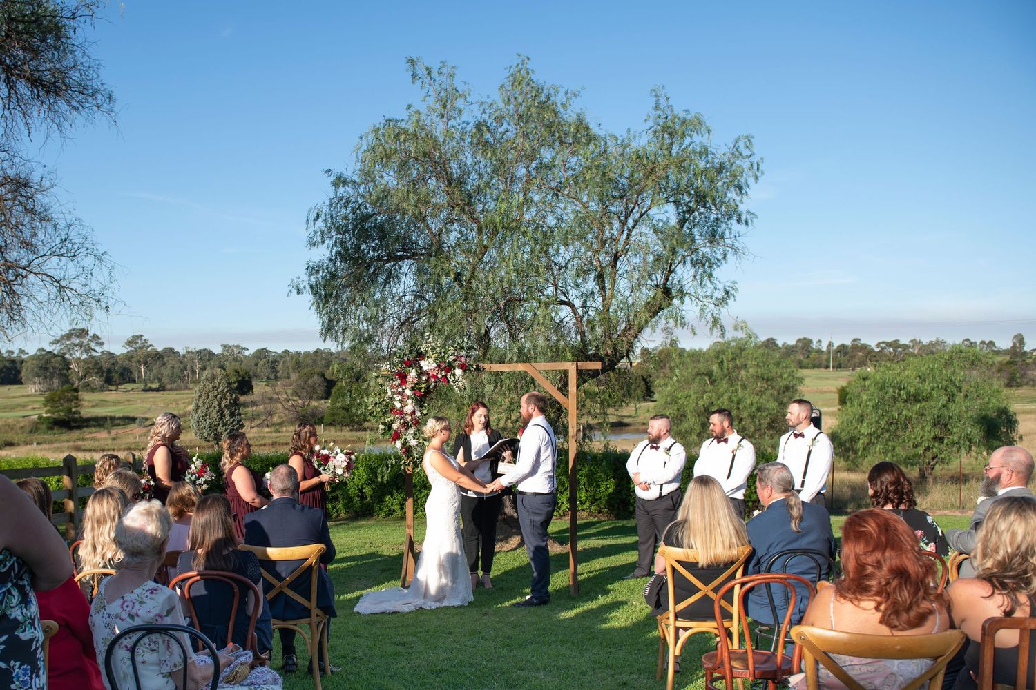 Guests gather outdoors for a scenic wedding ceremony under blue skies with trees in the background.
