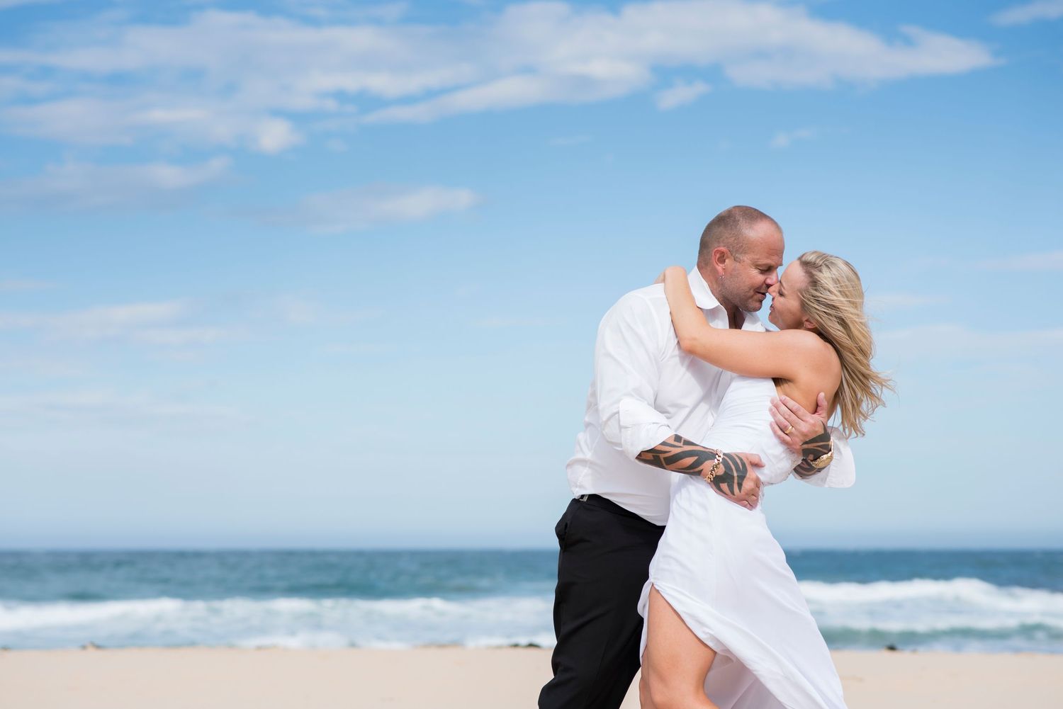 Bride and groom shares a romantic moment on a sunny beach in Wollongong with ocean waves and blue sky in the background.
