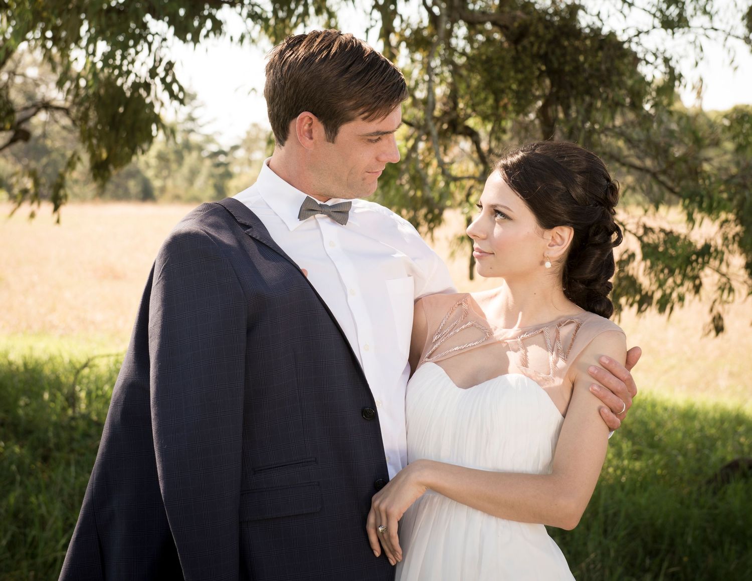 Romantic wedding couple embraces outdoors in a sunlit garden setting with trees in the background.