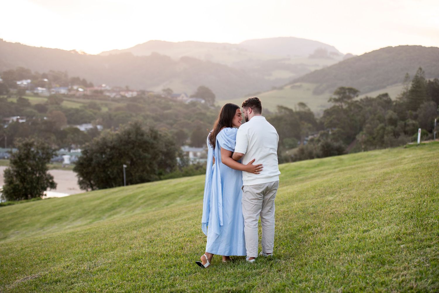 Romantic couple sits together on a grassy hillside during golden hour.