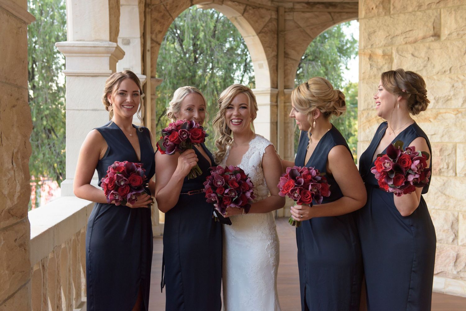 Bride with Bridesmaids in navy dresses and red bouquets poses together under a stone archway.