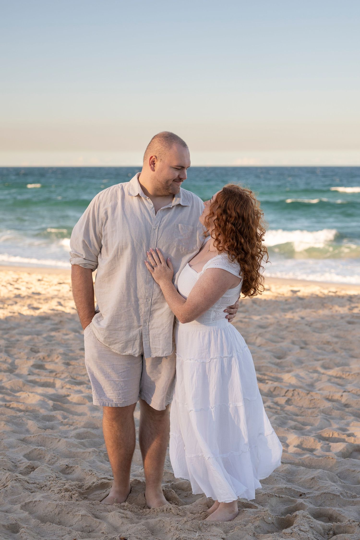 Couple in white outfits sharing a romantic moment on a sandy beach at sunset with ocean waves behind them.
