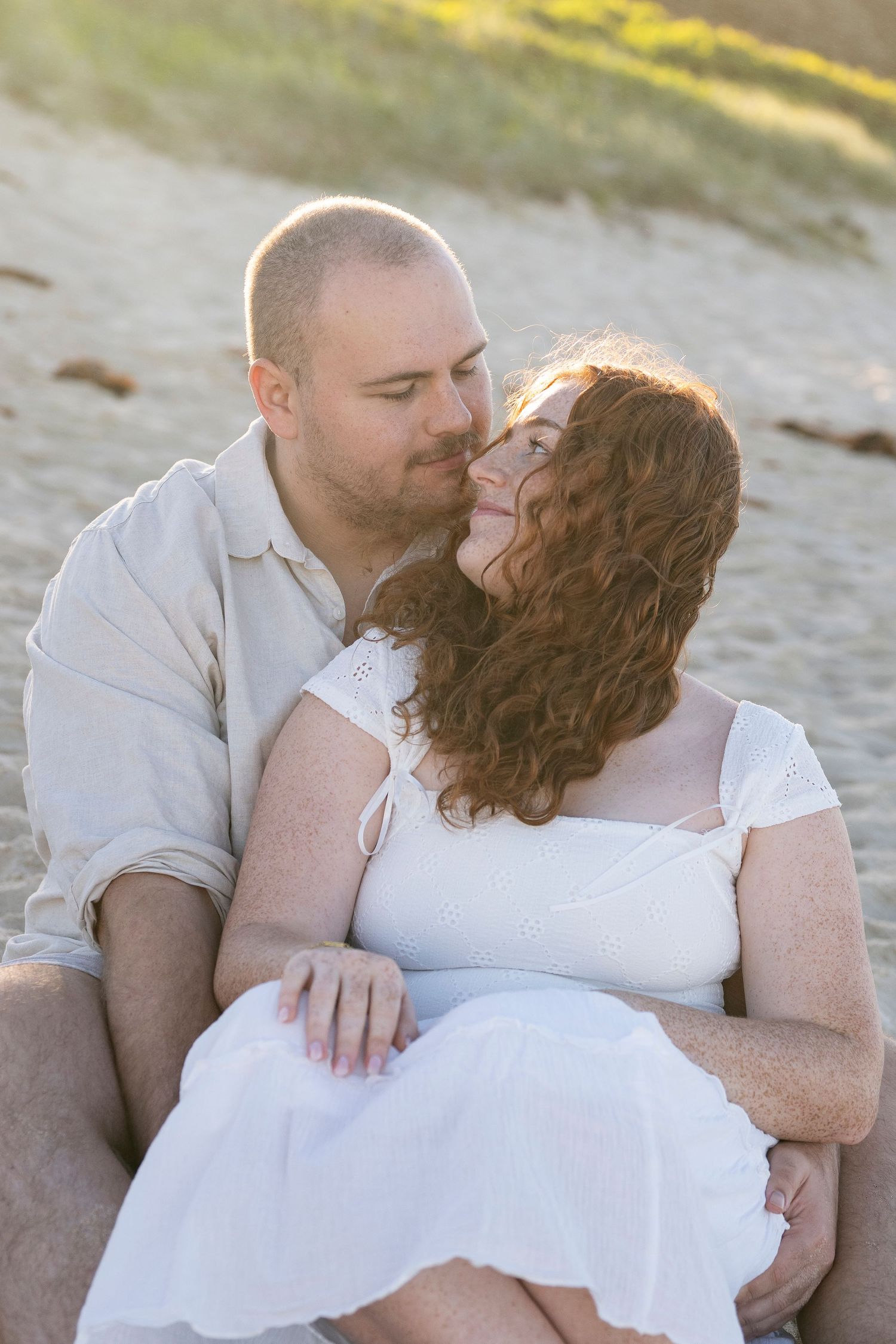 Romantic couple sitting together on a sandy beach, woman in white dress leaning against man in warm golden sunlight.