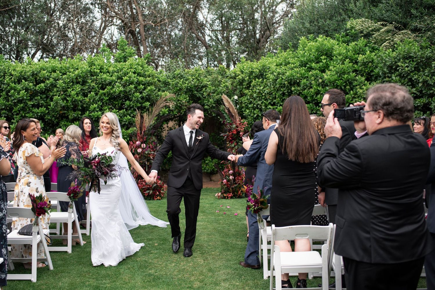 Wedding guests celebrate during an outdoor garden ceremony with raised hands and joyful expressions.