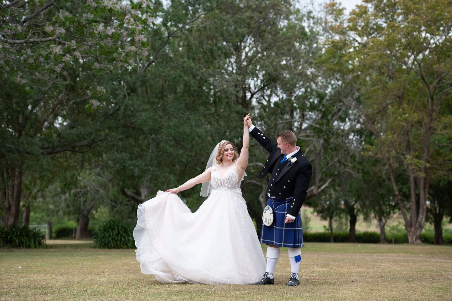 Bride in white gown and groom in Scottish kilt dancing joyfully outdoors in a lush green park on their wedding day.