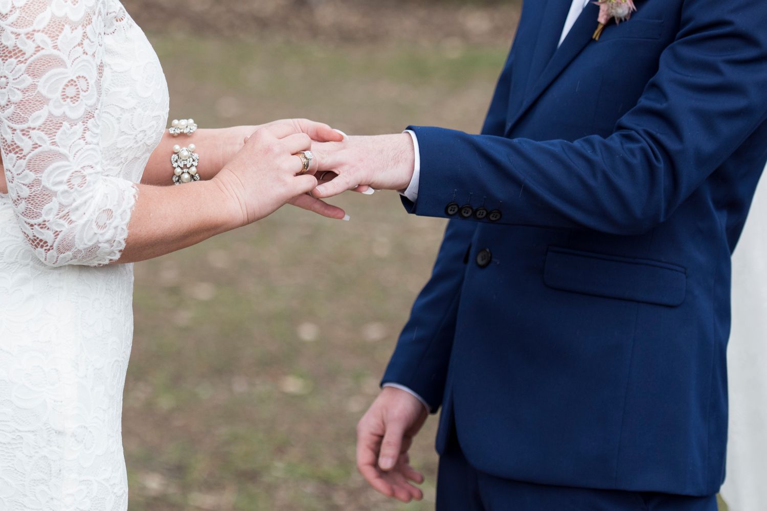 Close-up of hands exchanging wedding rings during ceremony with white lace dress and navy blue suit visible.