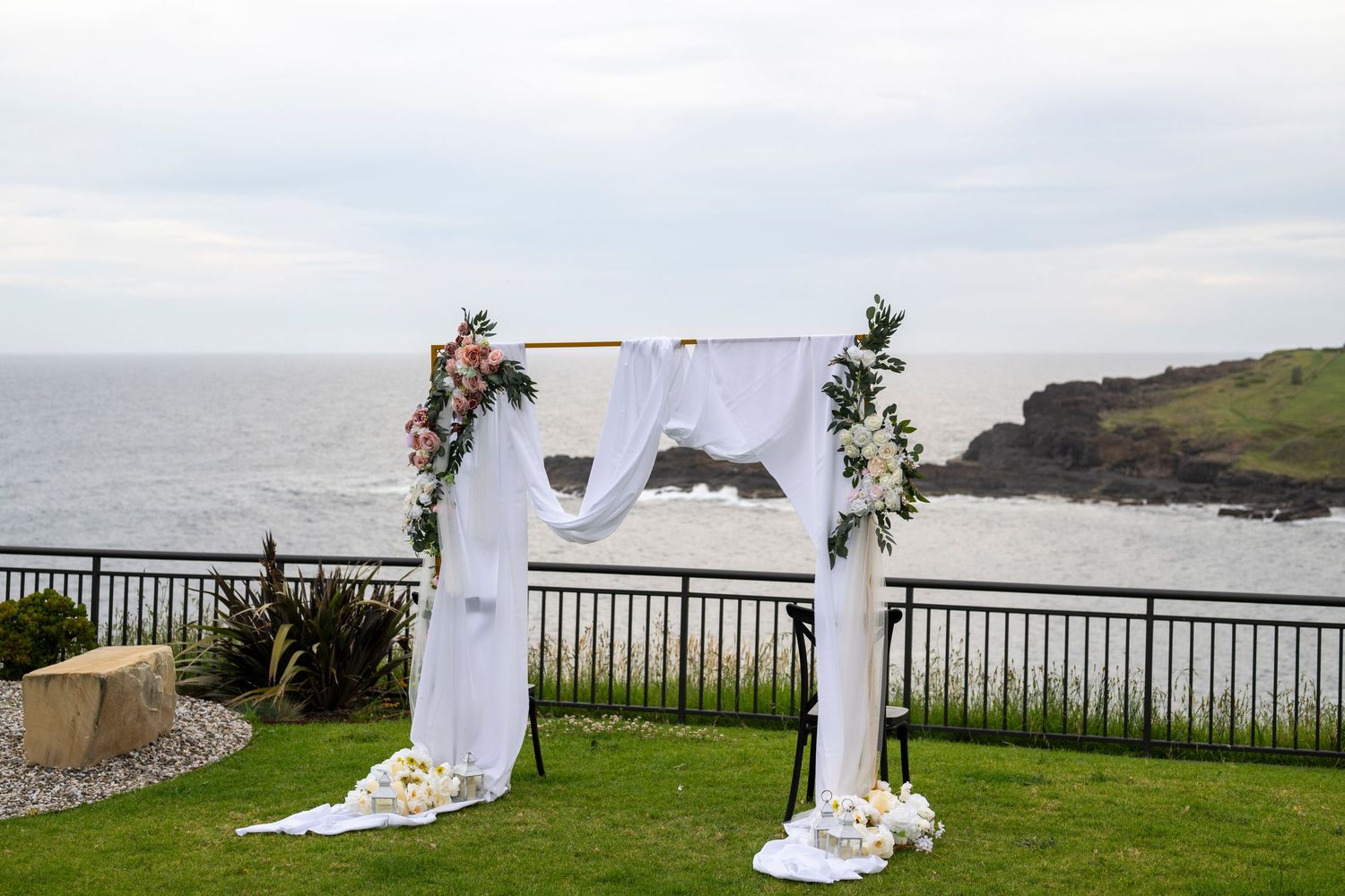 A white wedding arch decorated with floral arrangements stands on a clifftop overlooking the ocean on a cloudy day.