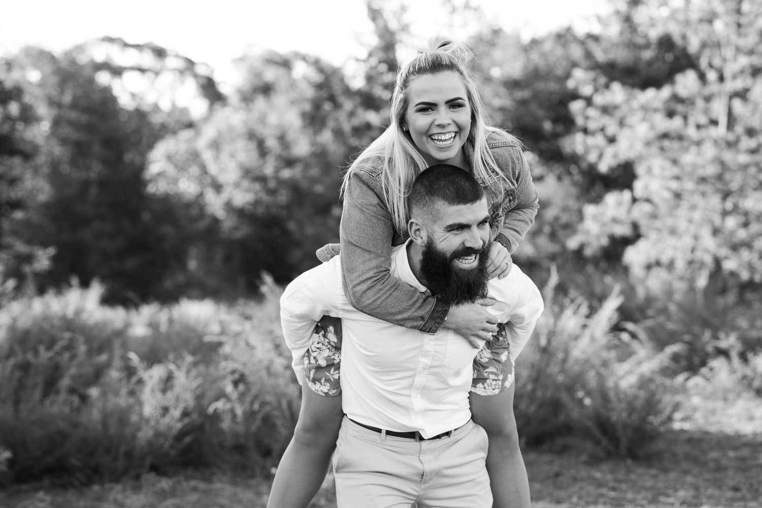 A playful couple shares a piggyback ride moment outdoors in black and white photography.