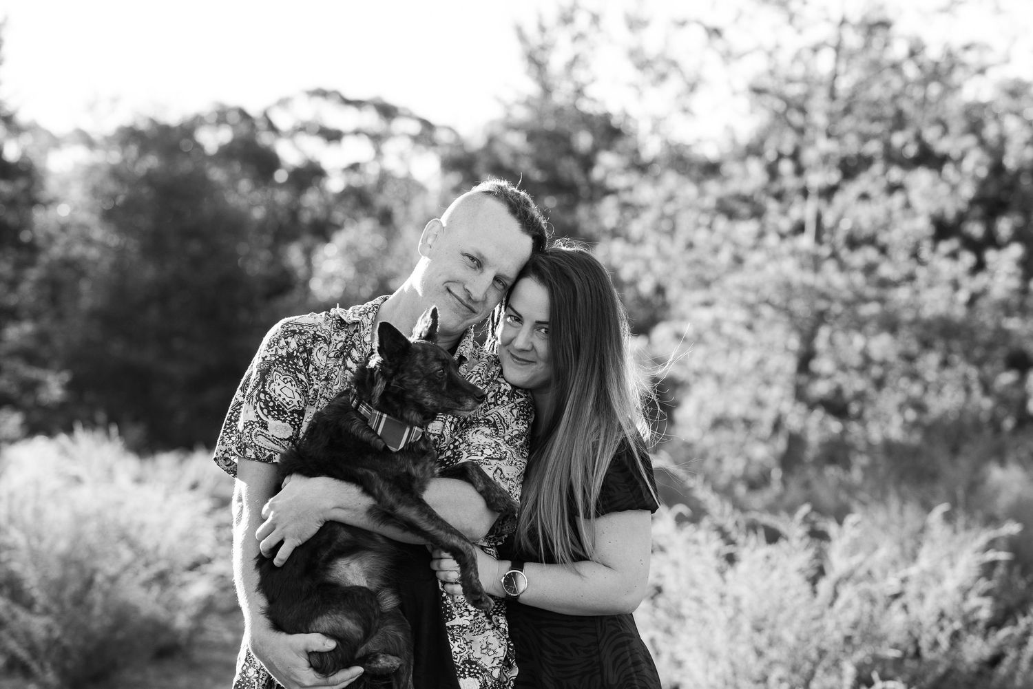 Black and white photo of a couple embracing in a natural outdoor setting with their dog.