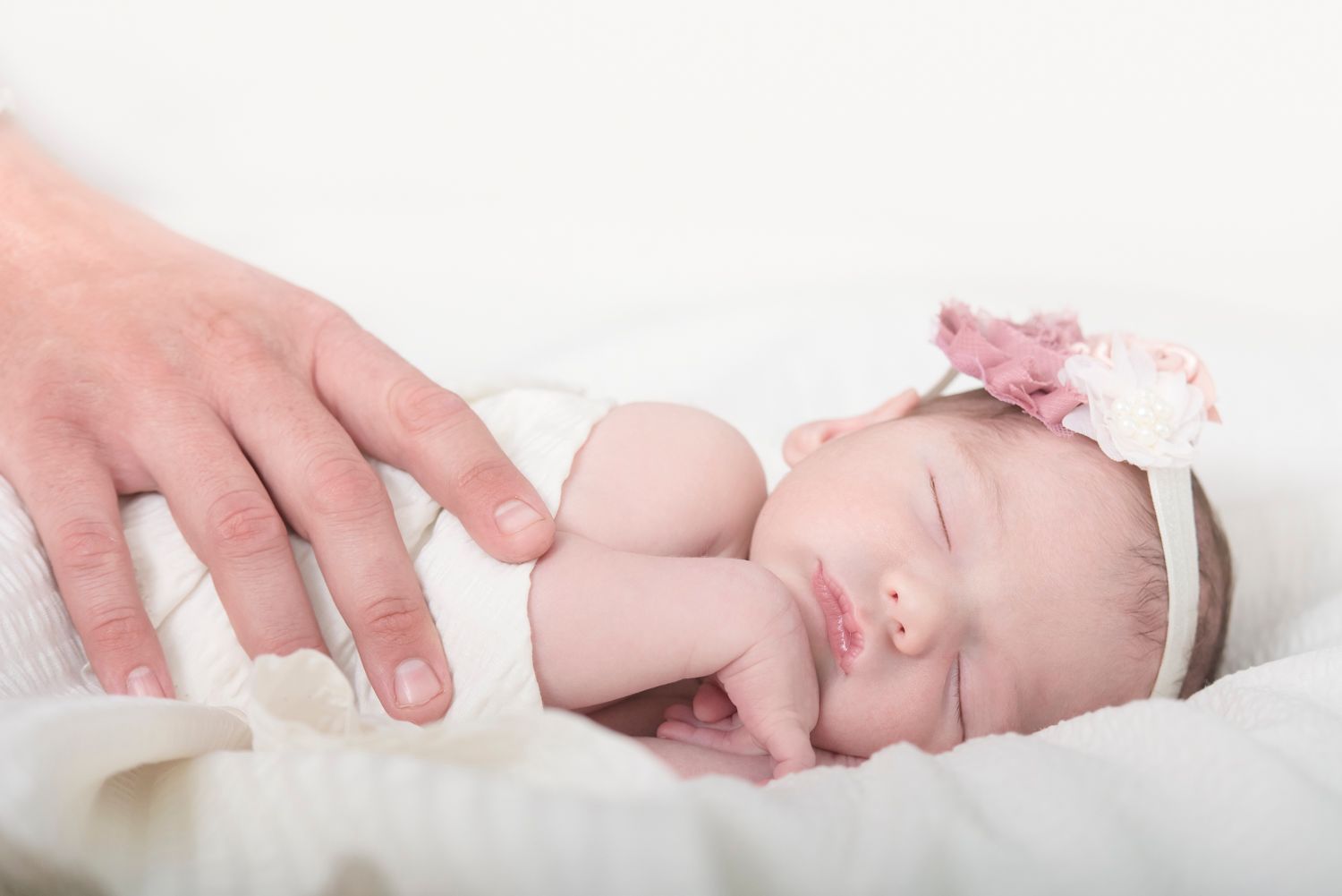 Sleeping newborn wrapped in white blanket with gentle hands touching.