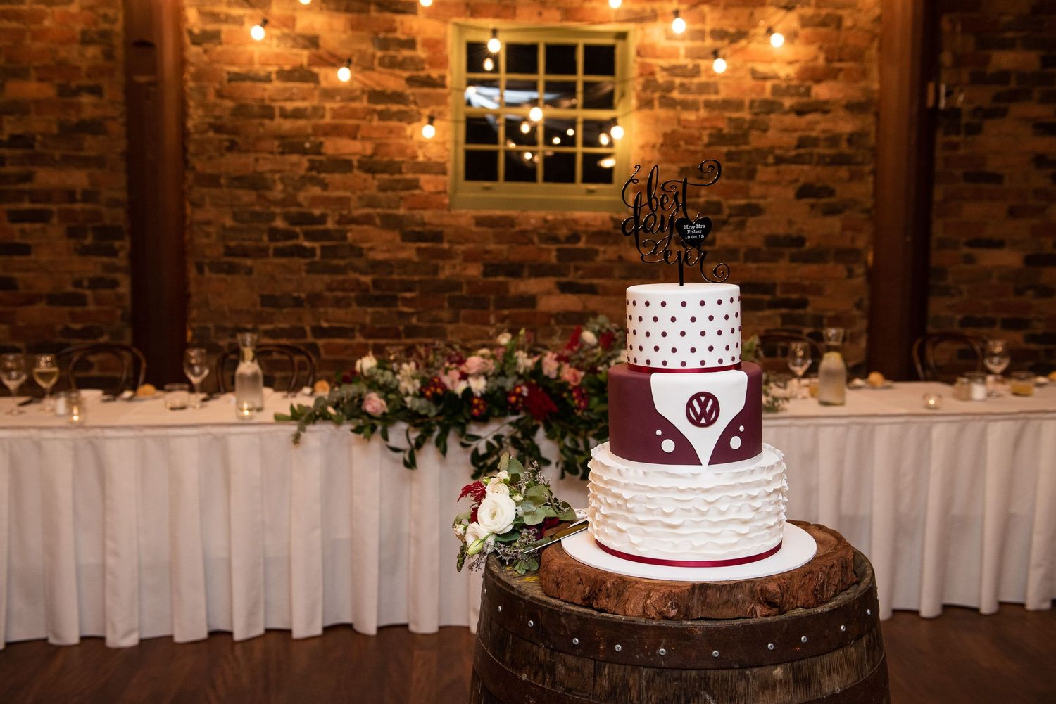 Elegant three-tier wedding cake with burgundy and white design displayed on wooden barrel in rustic brick venue.