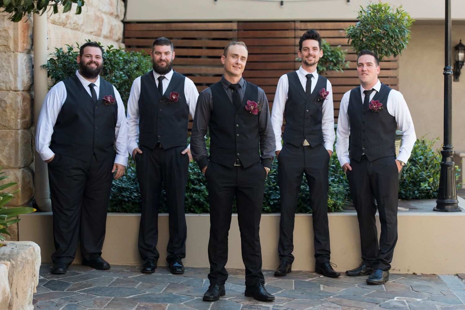 Groom with Groomsmen in black suits and white shirts stand in a line against a stone wall at an outdoor venue.