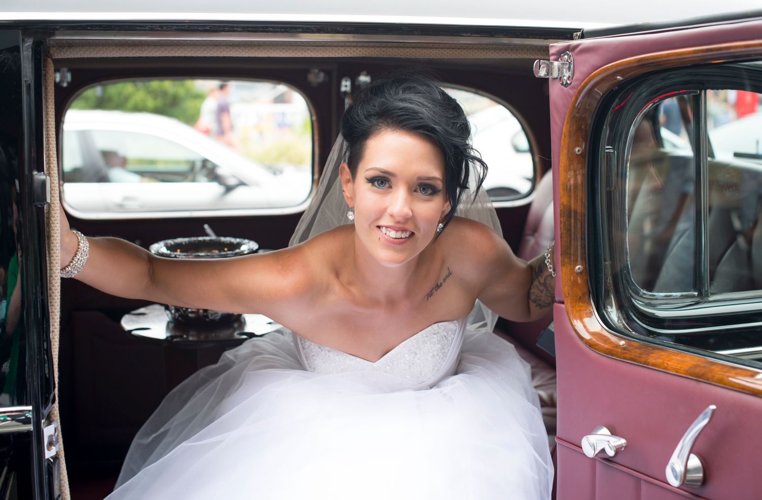 A bride wearing a white wedding dress sits in a vintage burgundy car on her special day.