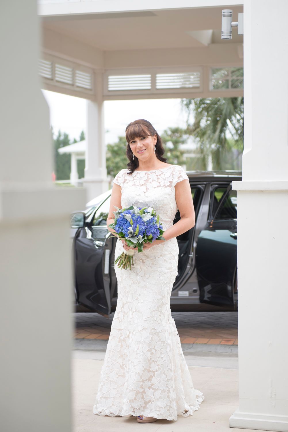 A bride in a lace mermaid wedding dress holds a blue floral bouquet while standing next to a black car.
