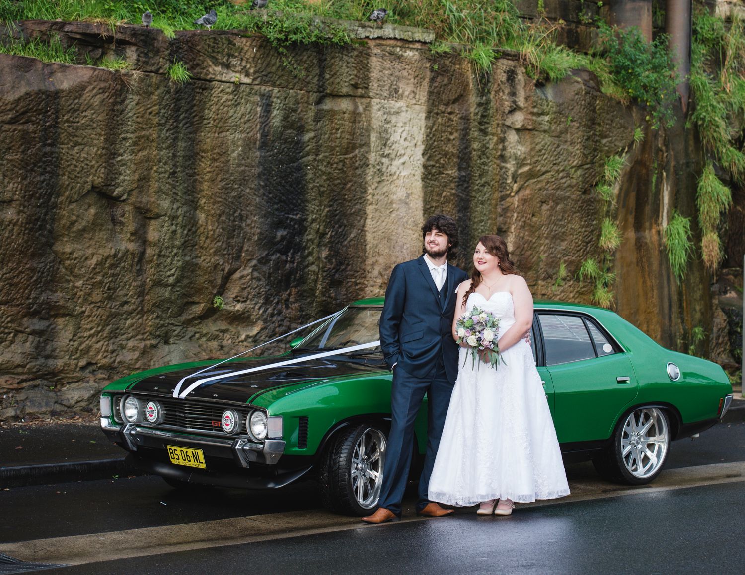 A wedding couple poses next to a classic green car against a stone wall in an urban setting.