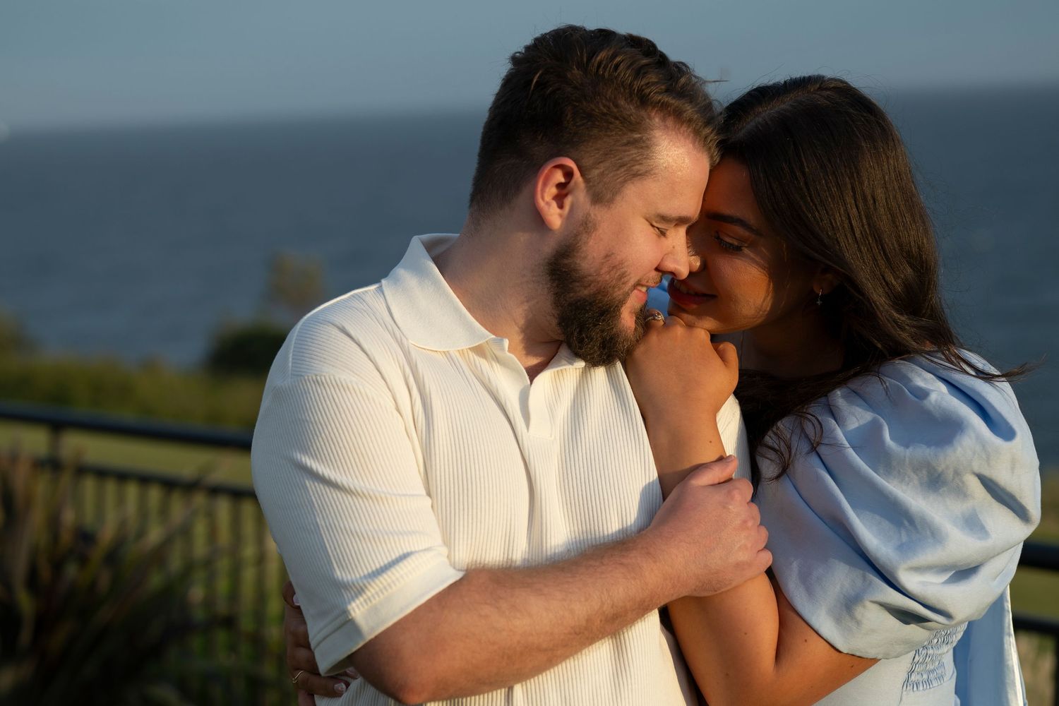 A romantic couple shares an intimate moment at sunset by the ocean, with warm golden light illuminating their embrace.