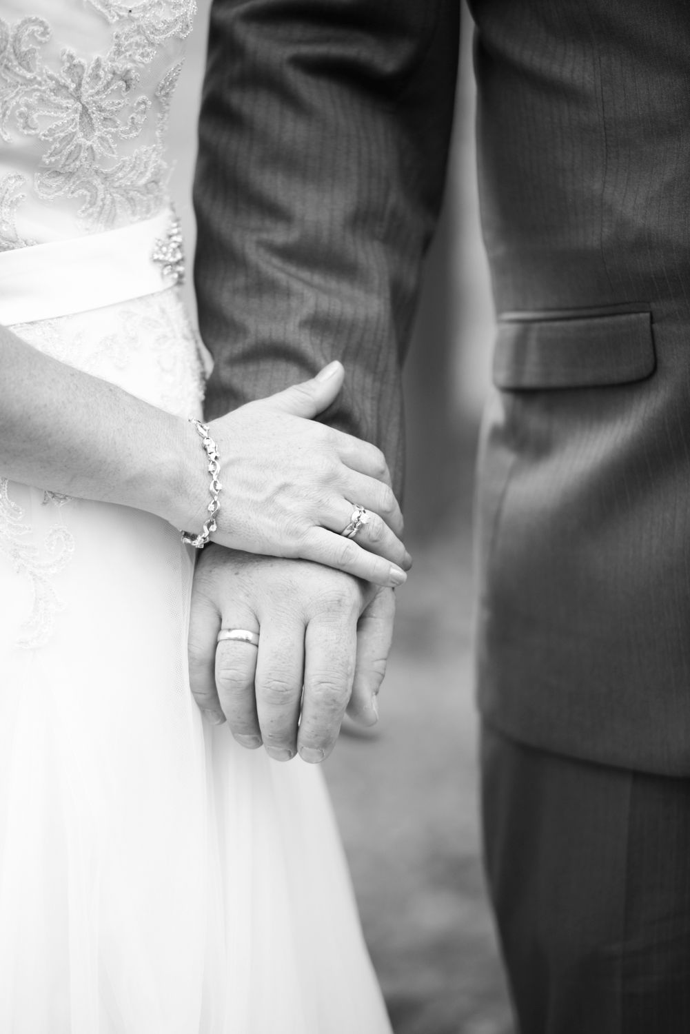 Close-up shot of newlyweds holding hands showing wedding rings and formal attire in black and white.