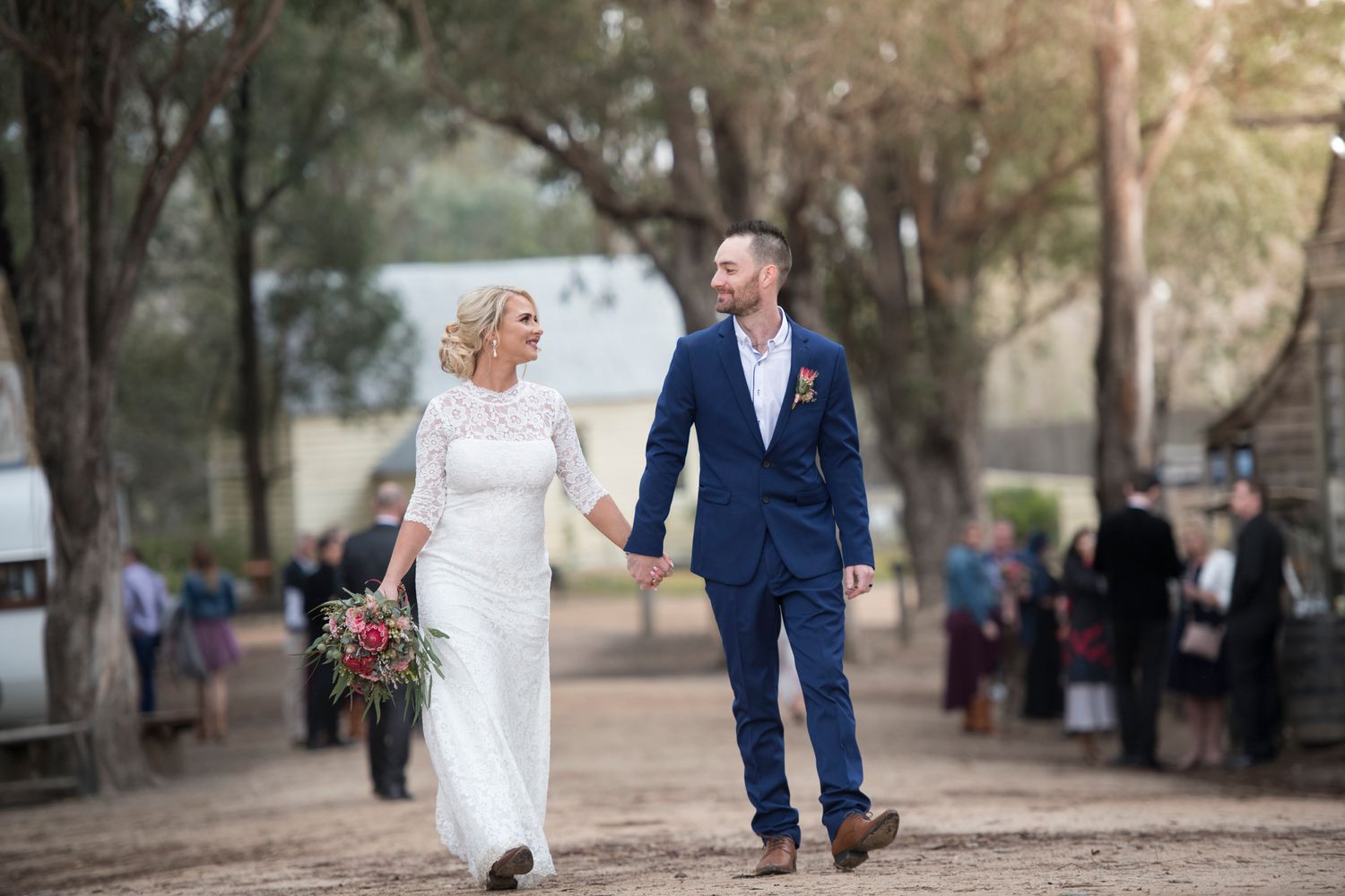 Bride and groom walks hand in hand along a tree-lined path on their wedding day with sunlight filtering through the trees.