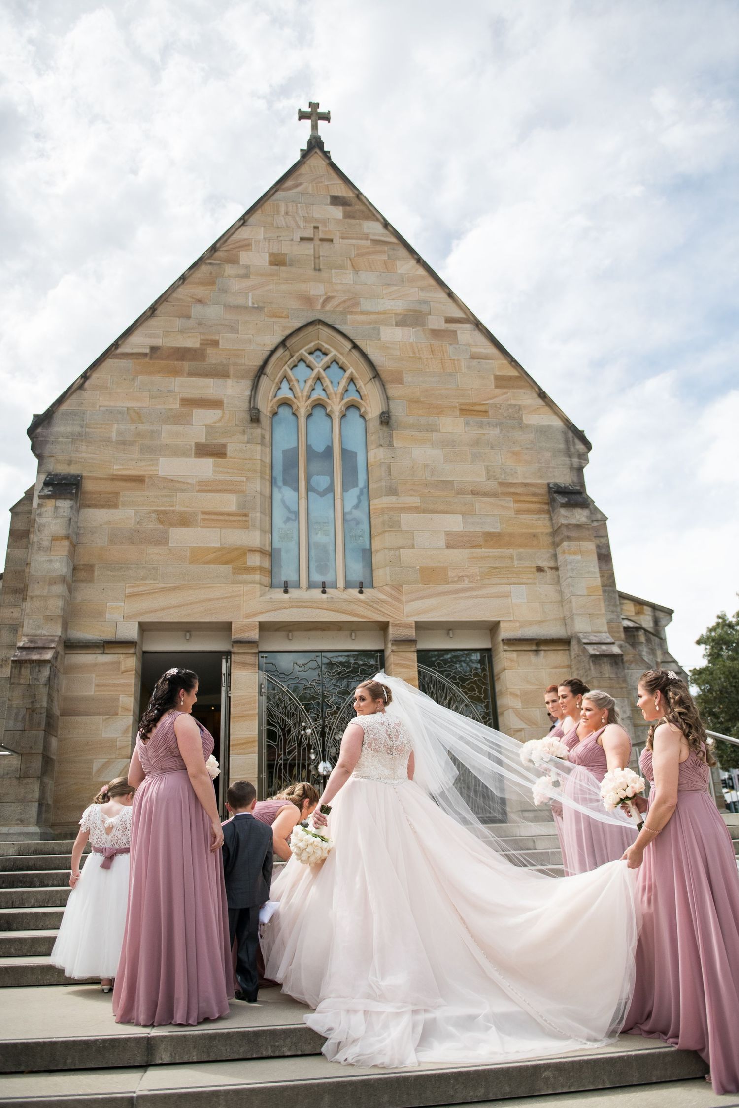 A wedding party poses outside a stone church with gothic windows as bridesmaids in pink dresses help with the bride's veil.