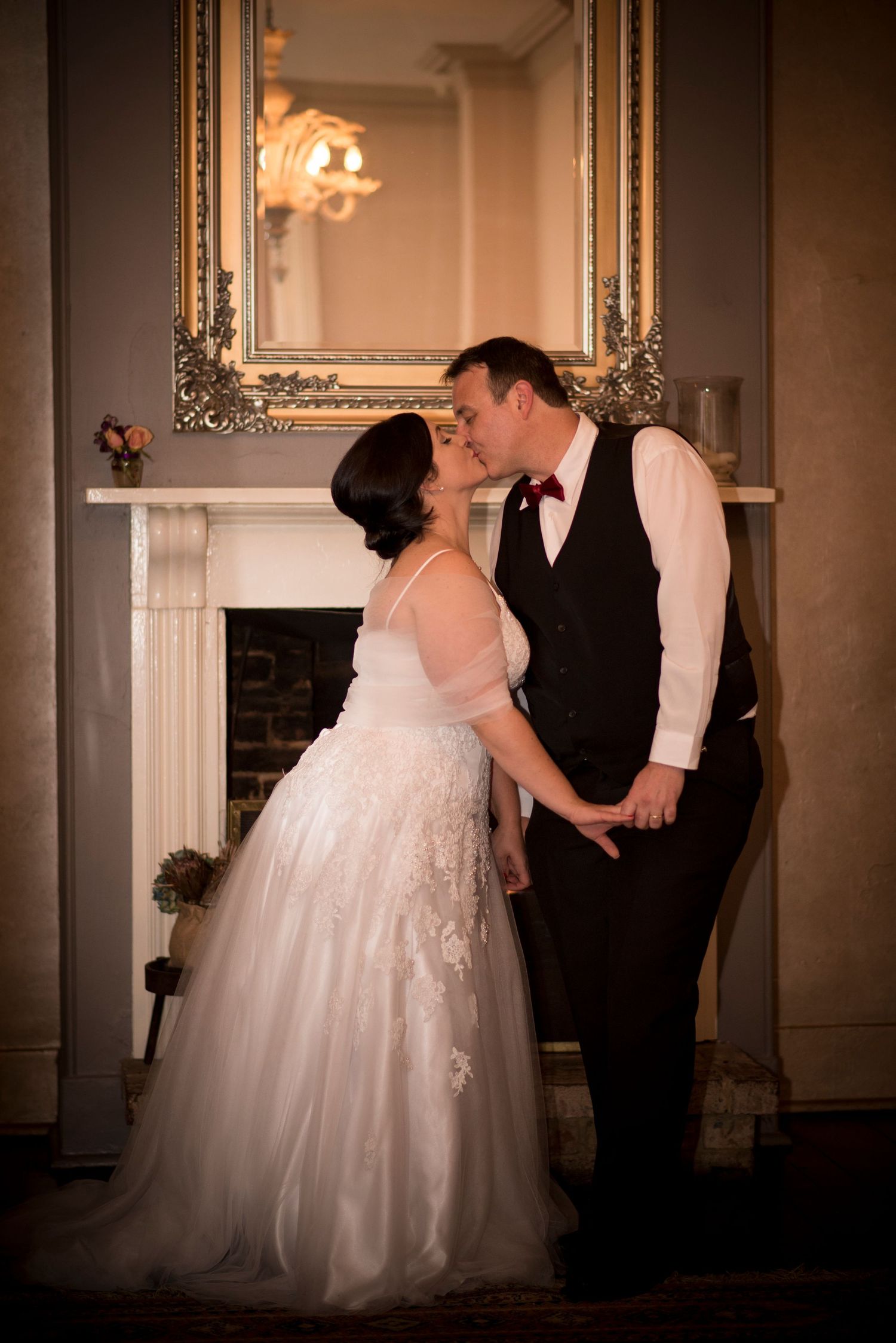 A romantic wedding couple shares a tender moment in front of an ornate fireplace with a gilded mirror.