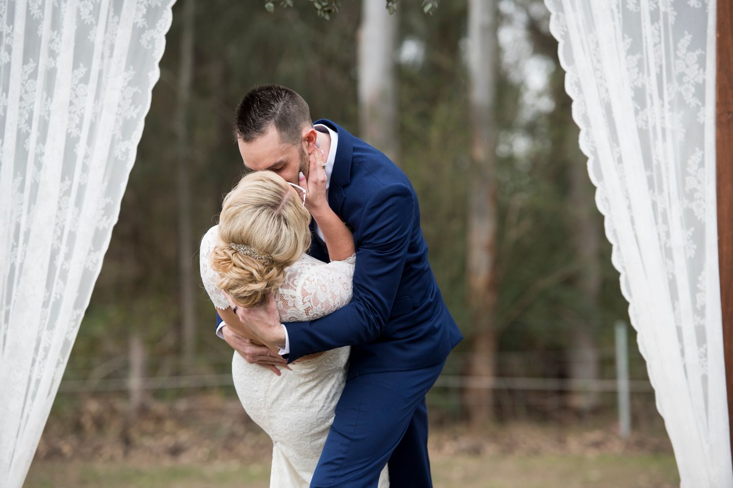 A romantic first kiss between newlyweds framed by white curtains in a bush setting.
