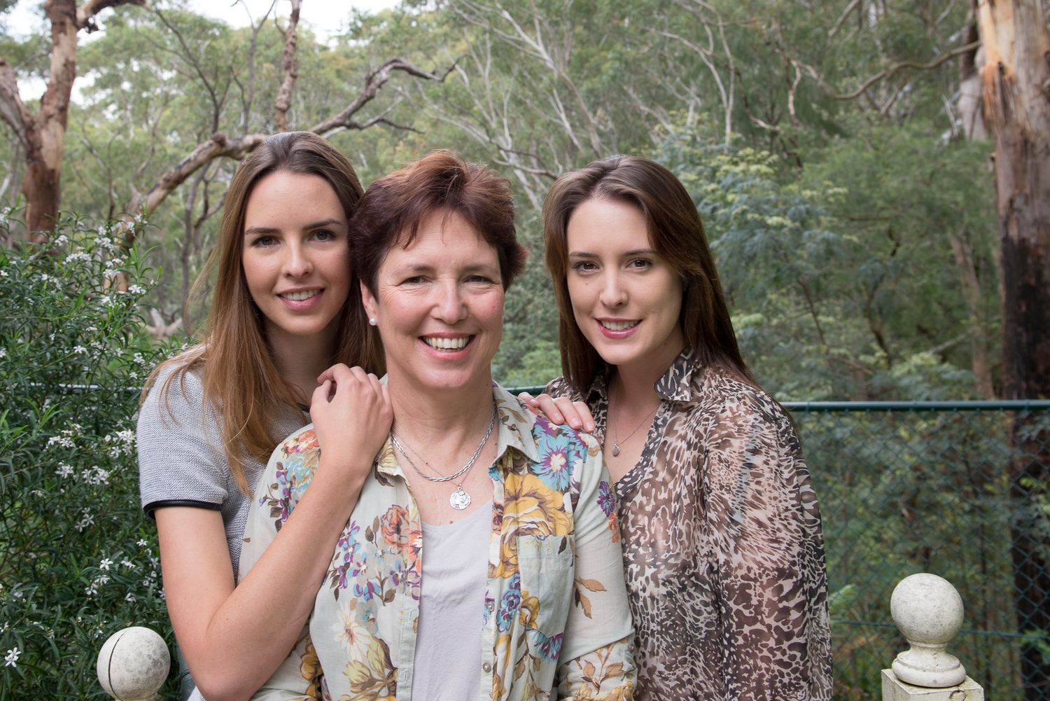 Mother and daughters smile and embrace during outdoor photo session in wooded setting.
