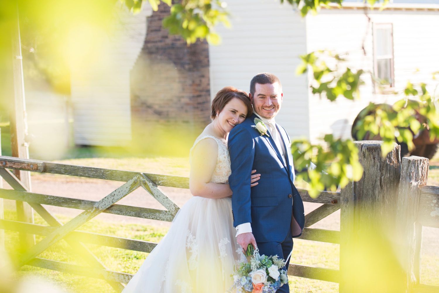 A couple in wedding attire embraces outdoors in a sunny garden setting with soft bokeh lighting effects.