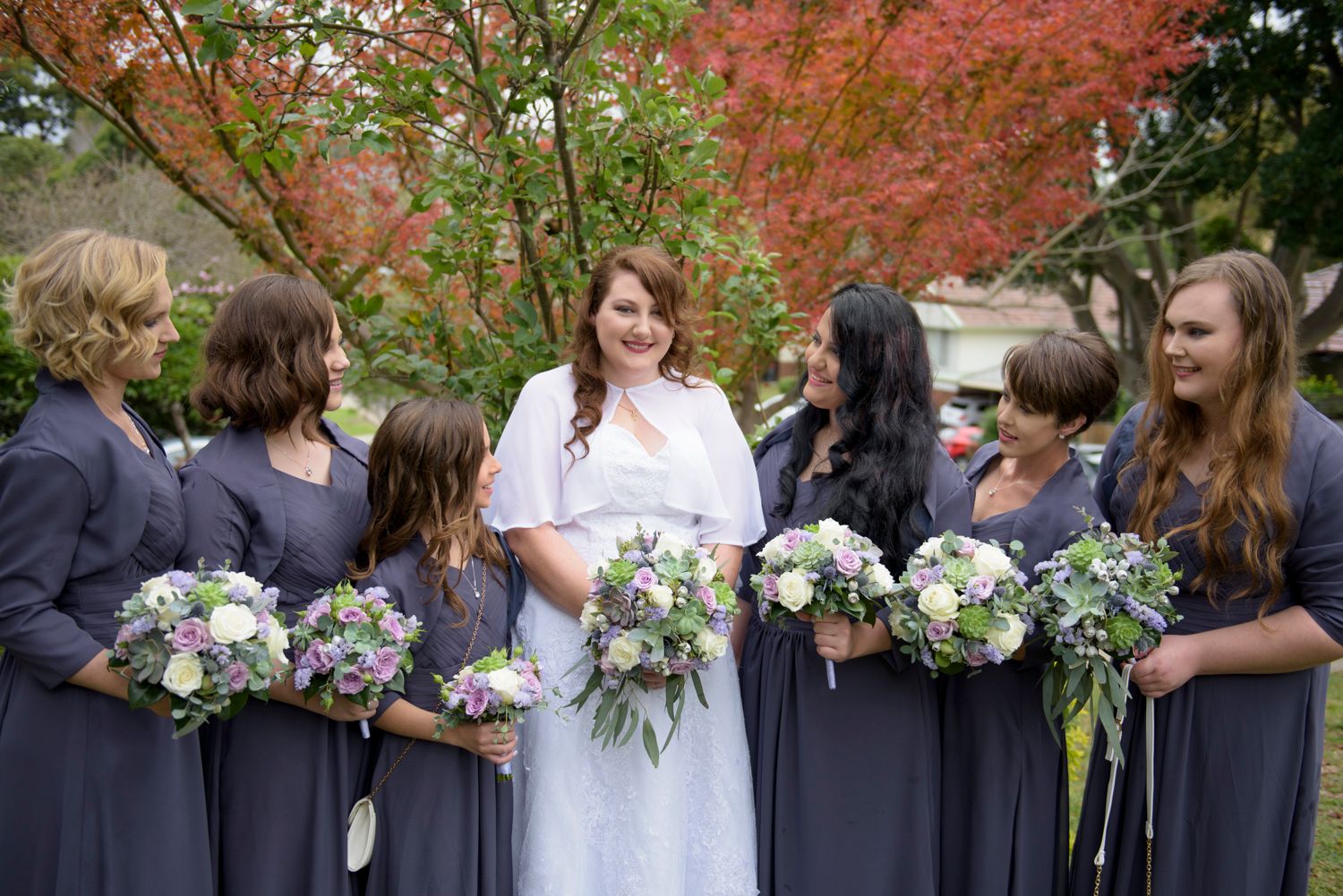 Wedding party poses together outdoors under vibrant autumn foliage in a garden setting.