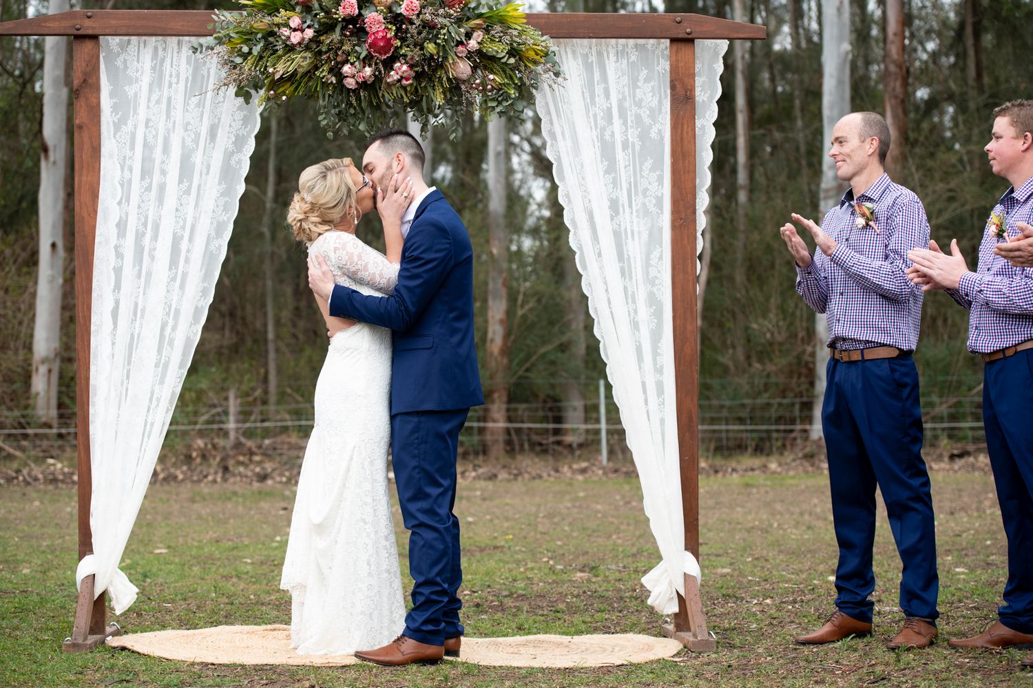 A couple shares their first kiss under a rustic wooden arch decorated with white curtains and floral arrangements outdoors.