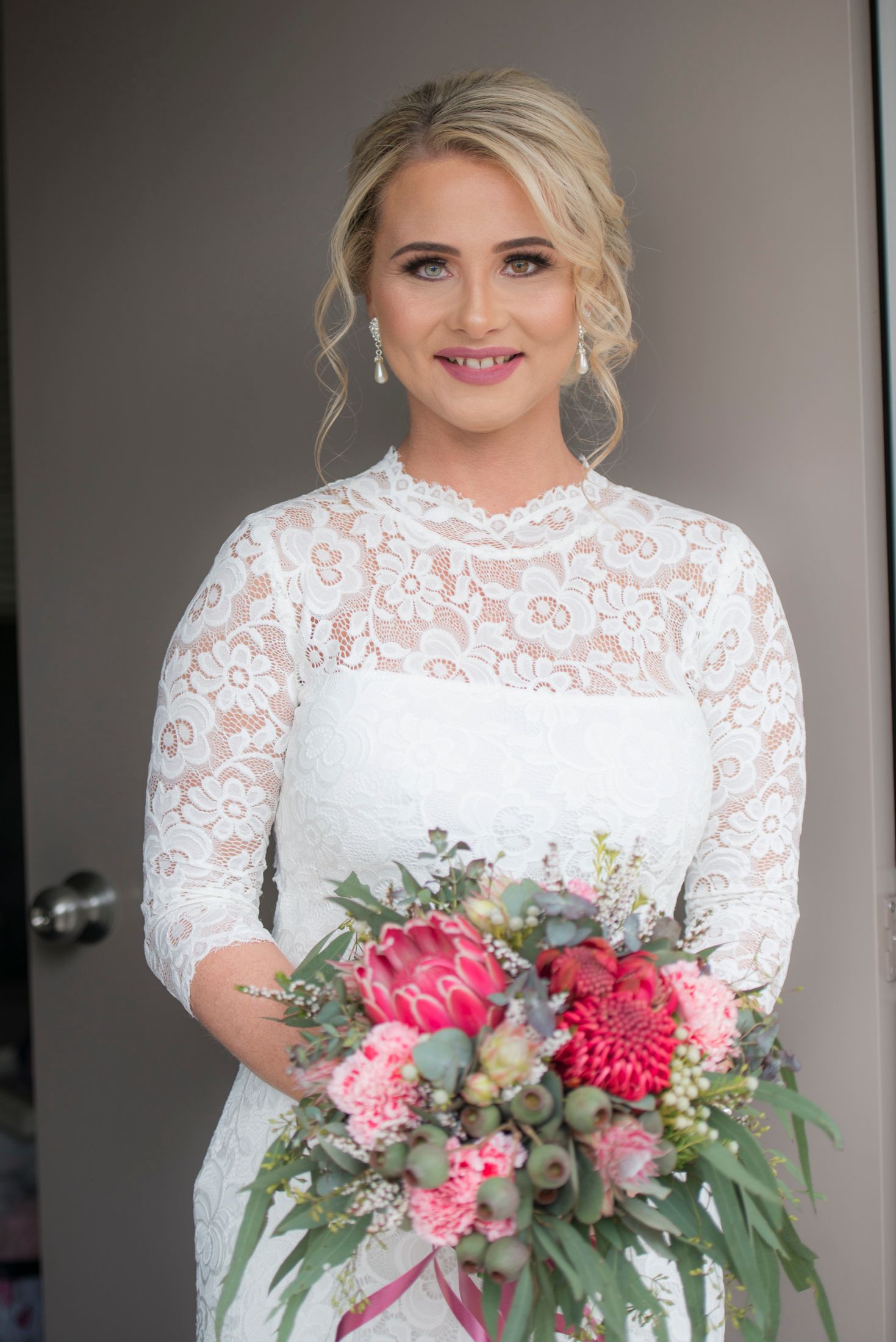 A bride in a white lace dress with long sleeves holds a vibrant pink and green floral native bouquet.
