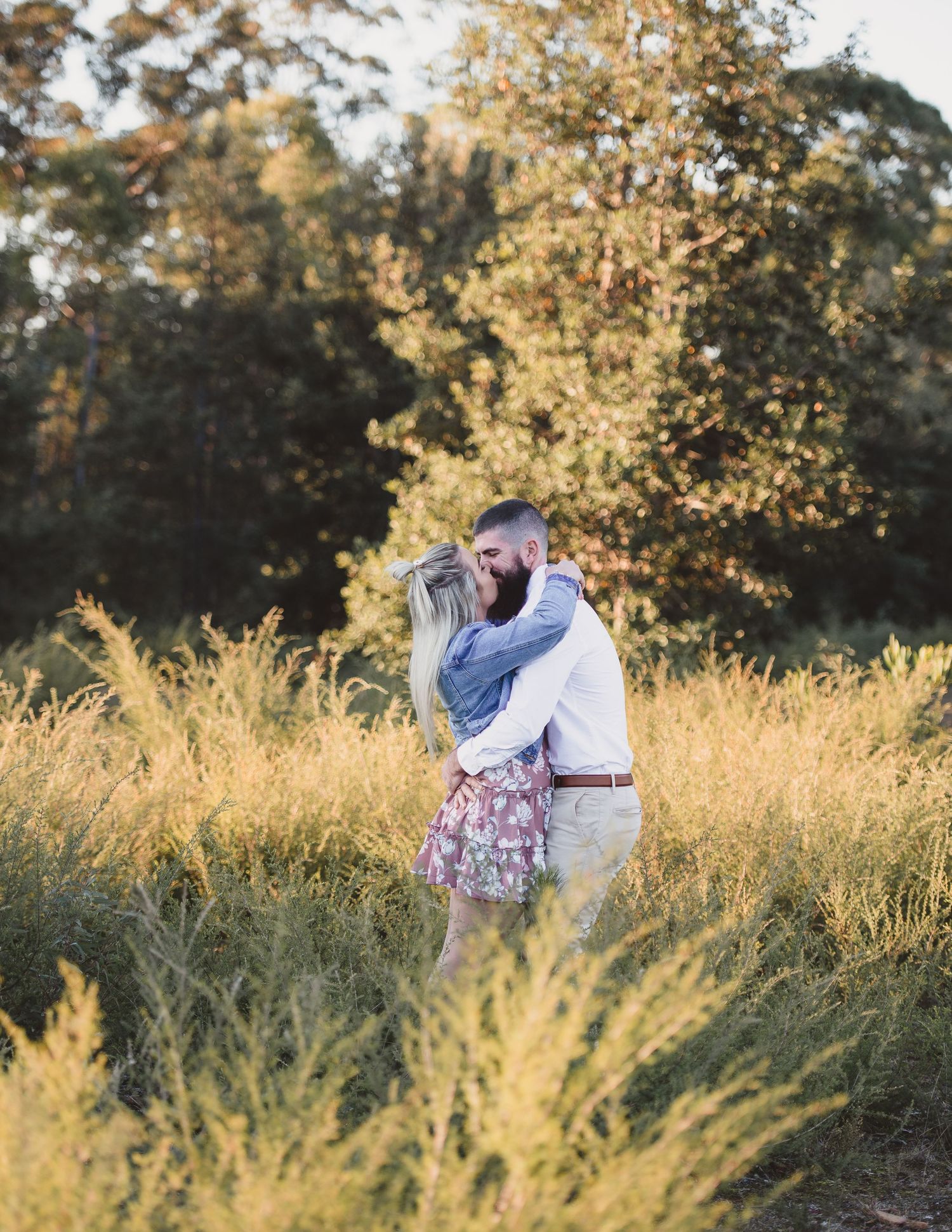 Couple embracing in a golden field during sunset with trees in the background.