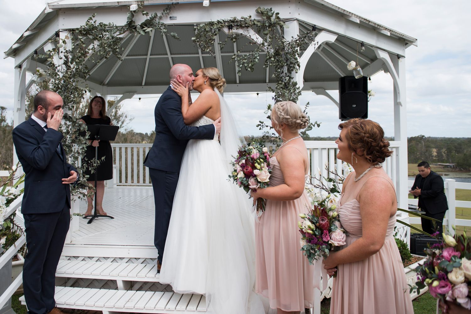 A romantic outdoor wedding ceremony takes place under a white gazebo decorated with floral arrangements.