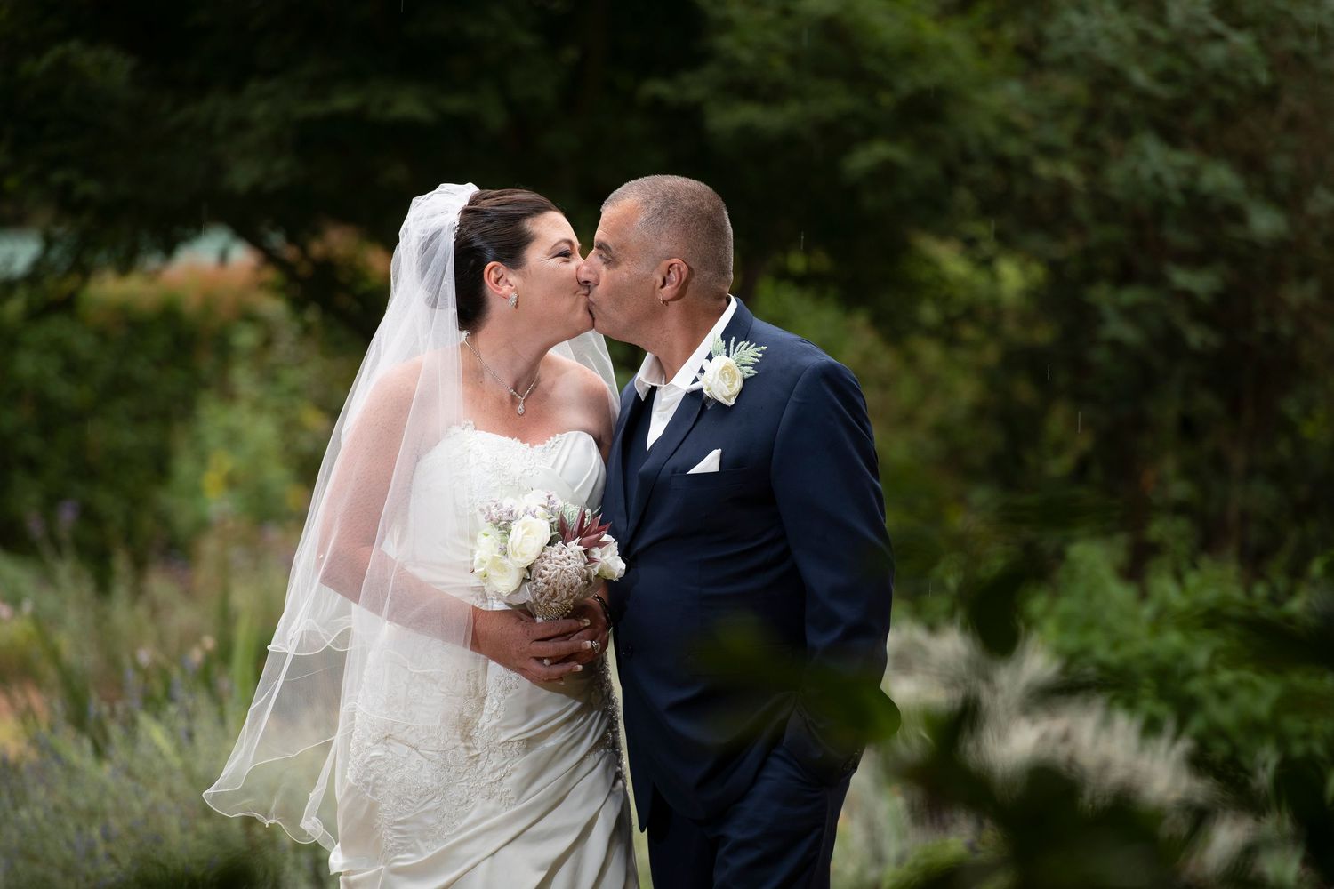 A bride and groom share a romantic kiss in a garden setting while holding a white floral bouquet.