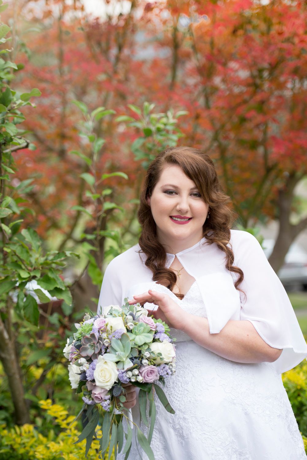 Bride in white dress poses with delicate bouquet against vibrant autumn foliage background.