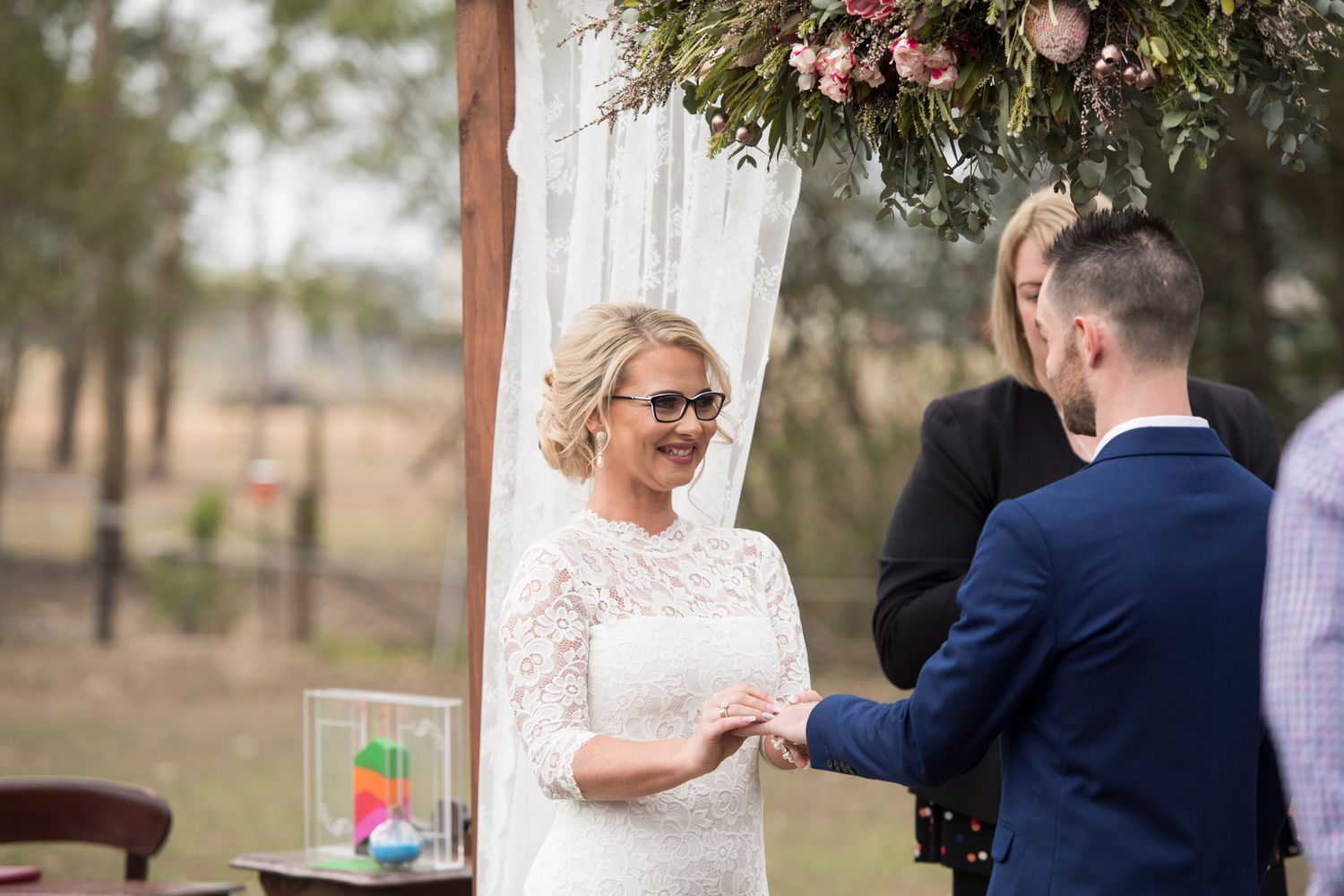 A romantic outdoor wedding ceremony features a couple exchanging vows beneath a floral arch in a natural setting.