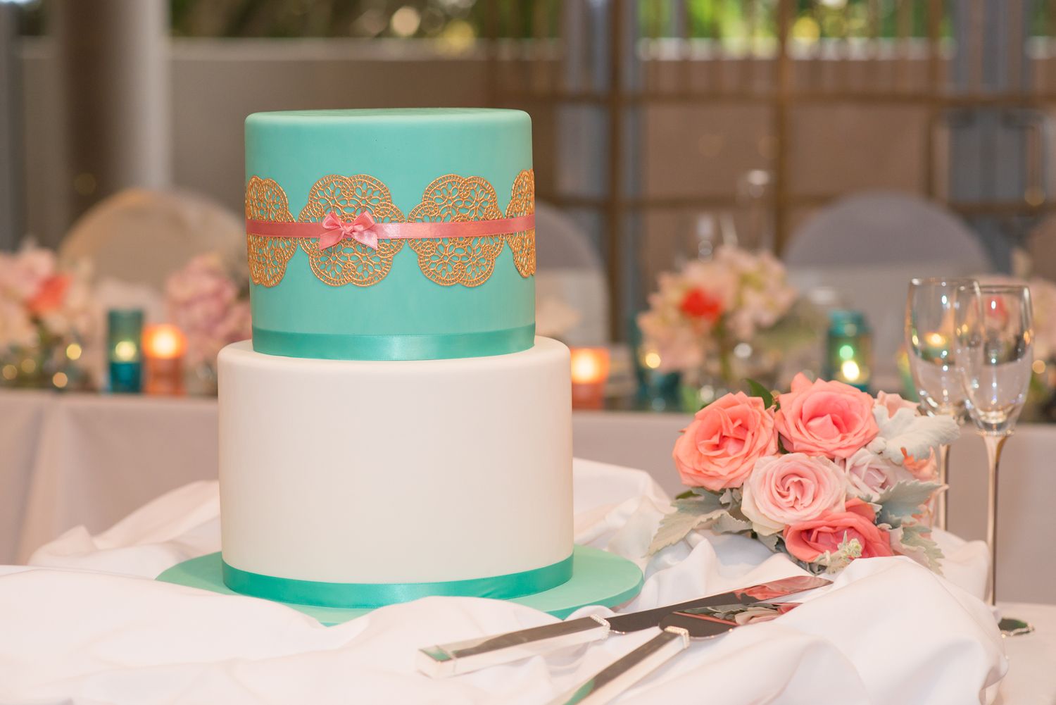 A two-tier wedding cake decorated in turquoise and white with coral lace ribbon, displayed with pink roses and table settings.