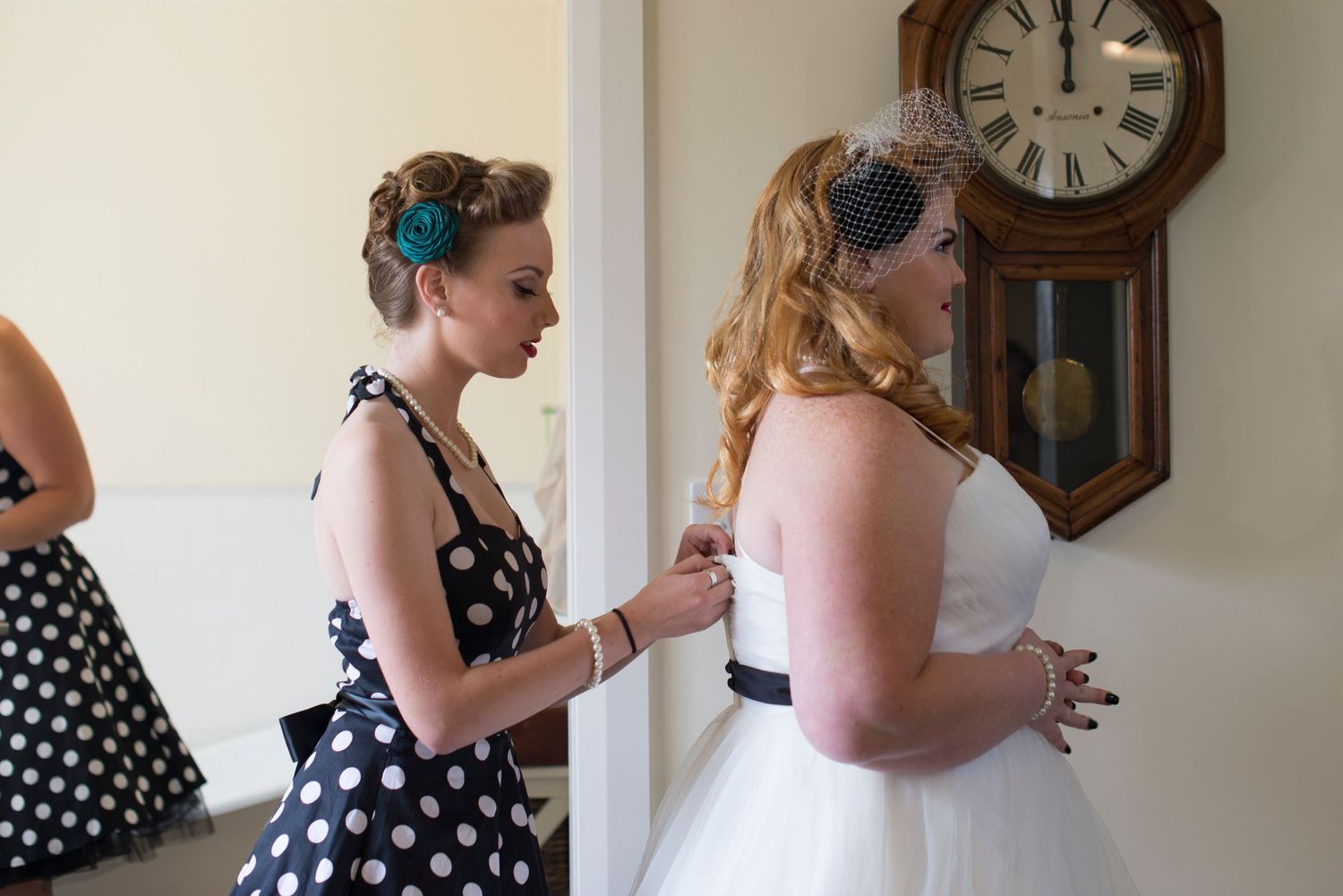 Bridesmaids in polka dot dresses help with wedding preparations in a room with vintage clocks on the wall.