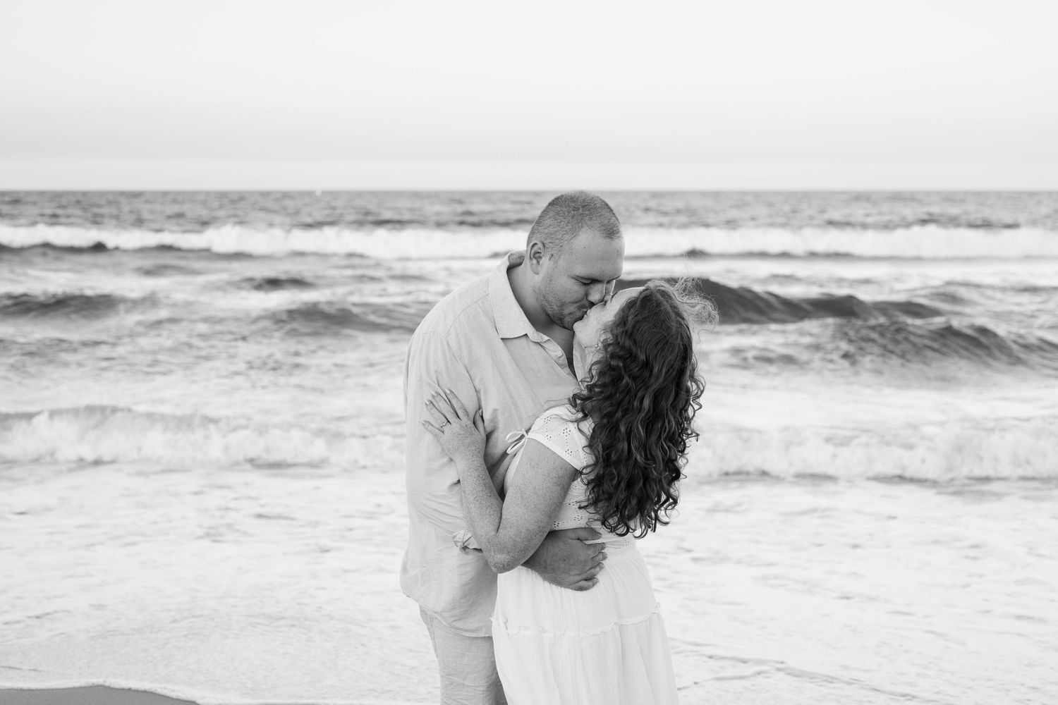 Black and white photo of a couple sharing a romantic kiss on the beach with ocean waves in the background.