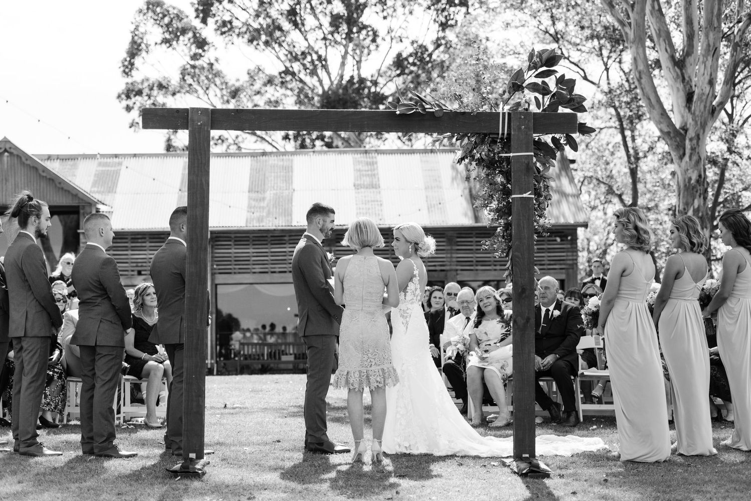Black and white photo of an outdoor wedding ceremony taking place under a wooden arch with guests gathered around.