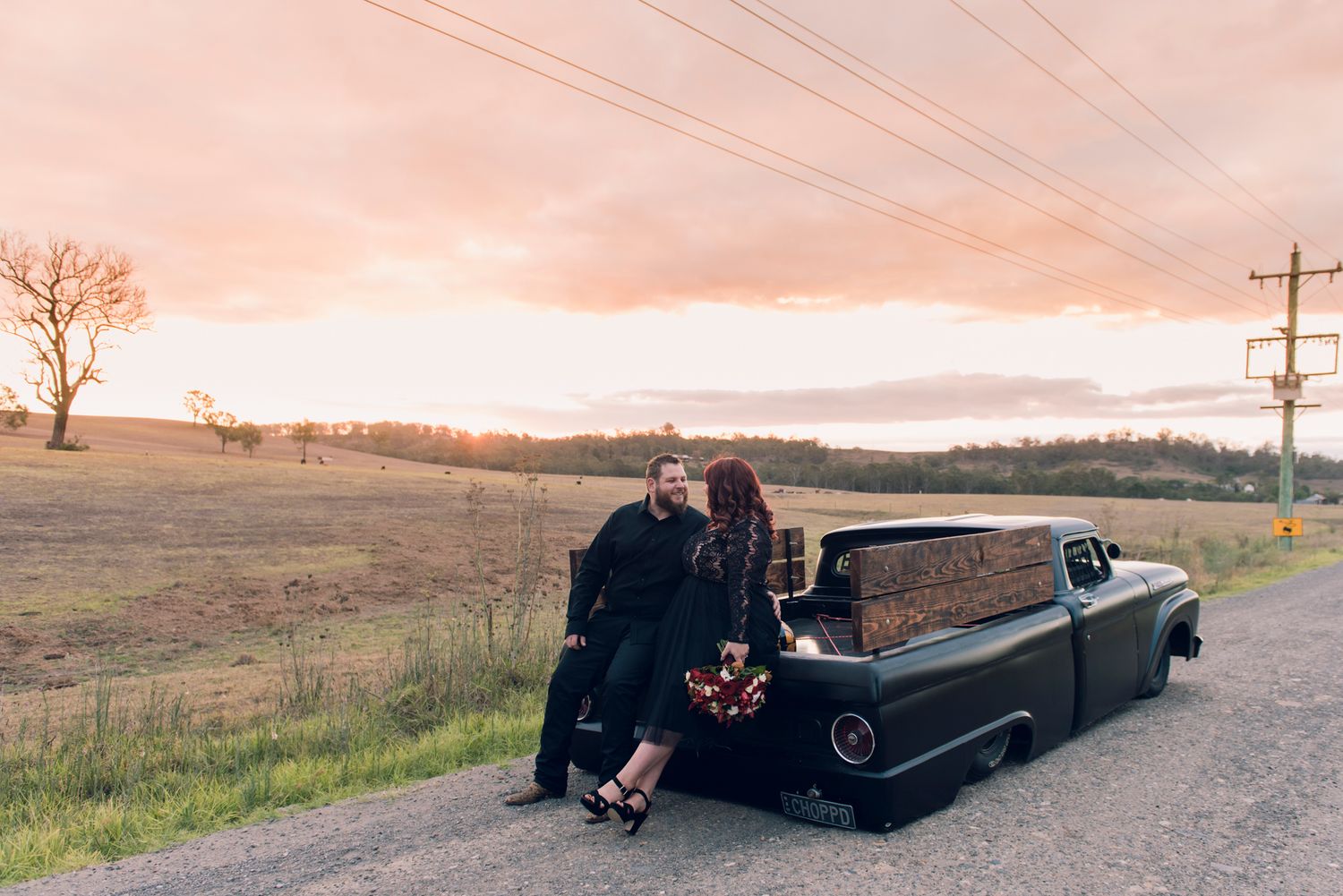 A vintage pickup truck parked on a rural road at sunset with figures sitting on the tailgate.