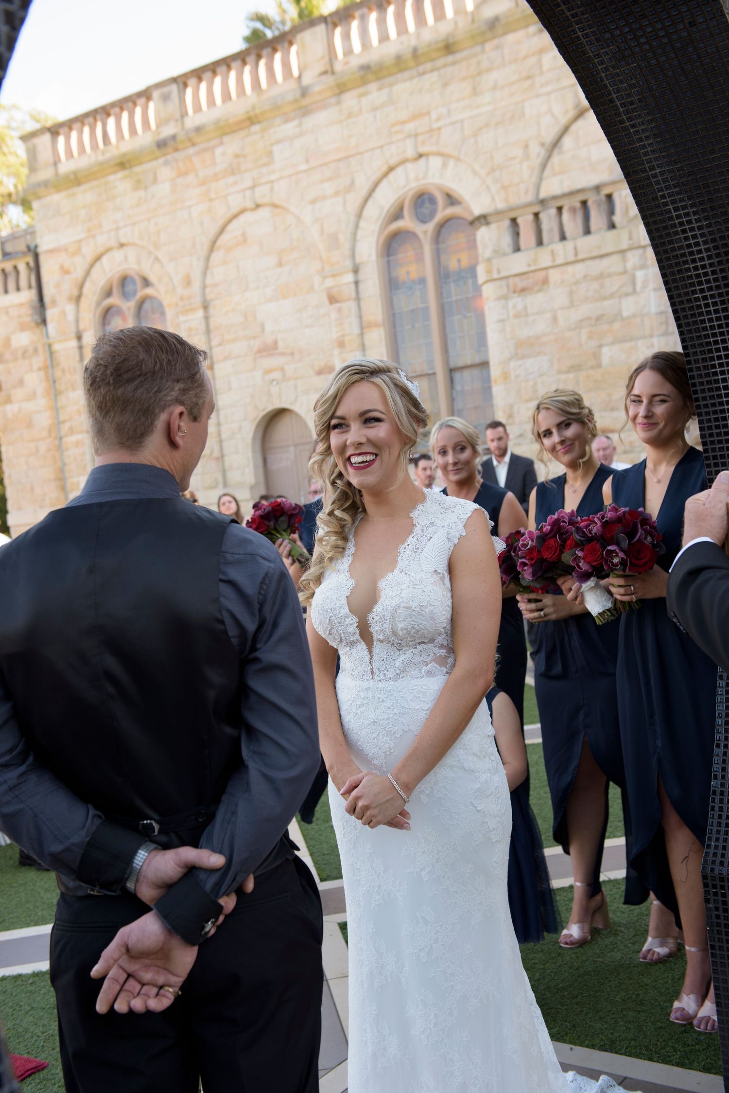 Wedding party gathered outside a stone building with red floral bouquets.